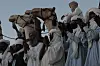 Men in Al Dein, Darfur, are wearing white and blue jalaib and turbans, clapping and chanting. Some are standing on the ground, and some are on camels.