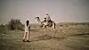 Abir Ibrahim is standing in the desert, speaking to a man wearing a white turban on a camel.