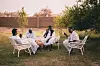 Four men in white jalaib and black wests are sitting on two white chairs and one angareeb in a field, drinking tea and eating legemat.