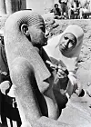 Black and white picture of a woman wearing a white hijab, brushing an ancient Egyptian statue of a woman. Both share the same features of a round face and a straight, wide nose.