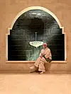 Eman Onsy, wearing all white and a white and green cardigan, is sitting in an arched window that opens to a dark green fountain with an oriental lamp.