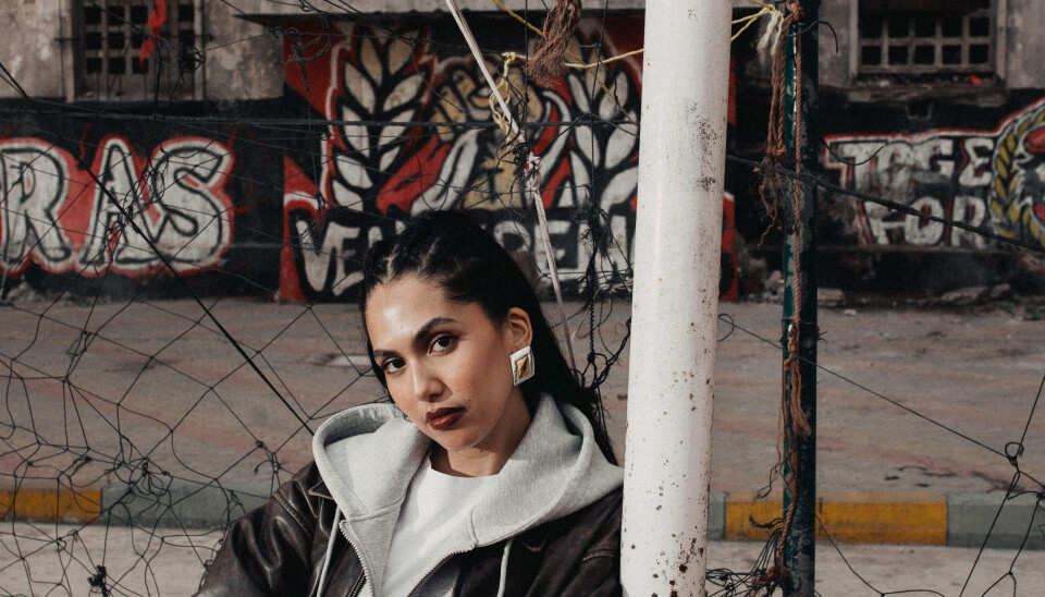“I always put myself into different skins, and I like to talk to everyone. You find me yapping everywhere all the time.” - Manal Benchlikha Manal is leaning on a goalpost, wearing a grey sweater and a black leather jacket, looking into the camera with a serious facial expression.