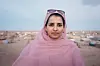 A woman wearing a pink toub is standing in the desert. Behind her are small houses that appear to be a refugee camp.
