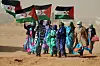A group of Sahrawi women in brightly colored clothes, holding up Western Saharan flags, waving in the wind.