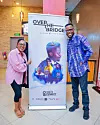 Tolu Ajayi, wearing a blue and purple patterned adire shirt, stands beside a guest in front of a promotional banner for his film Over the Bridge at a cinema venue.