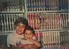 Malik, a young child, holds his little sibling, Sara, a toddler, in front of a shelf full of Disney VHS tapes.