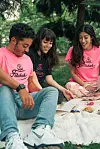 Malik, Sara, and Hana are sitting on a picnic blanket, looking through a notebook and smiling. They’re all wearing “Felukah” t-shirts, Hana wearing a black t-shirt with pink writing, and Malik and Sara wearing black t-shirts with pink writing.