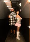 Khalid and Amira are standing in what appears to be the hallway of a nightclub. Khalid is wearing a black and white checkered shirt and a kuffiyah bandana, and Amira is wearing a pink mini-skirt and white stiletto boots.