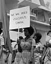 A group of women workers wearing khaki uniforms march past the Independence Arch in a May Day parade at the Black Star Square in May 1965. The woman in focus carries a placard reading, 'WE WILL BUILD OSAGYEFO'S GHANA'. Taken in 1965.