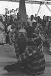 Two men in traditional attire perform, one with a drum while the other dances, in front of community members and elders at the Kodzidan. Renowned playwright Efua Sutherland sits on a step above the stage.