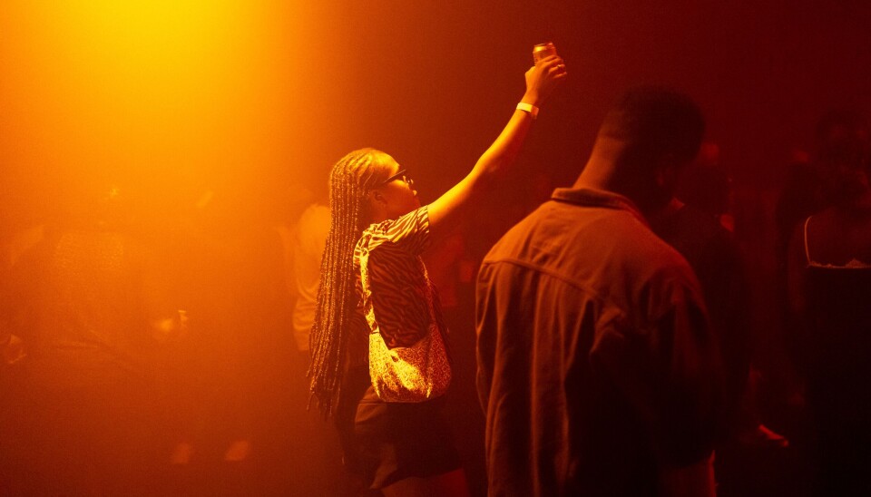 “For as long as I can remember, Detty December in Nigeria has always buzzed with a palpably boisterous energy — Nigerians are famously sanguine.” — Chibuzo Emmanuel. A woman dances, drink in hand and hand in the air, an orange hue around her and others near her.