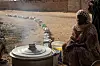 A woman wearing a toub is sitting next to a large pot and bucket. Behind her is a long line of smaller containers that are waiting to be filled with food.