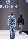 A model on the runway wearing Boyedoe. Her outfit is a blue patchwork denim coat-dress with a brown leather belt.