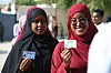 Two women in hijabs stand in line, holding up their ID cards. One wears glasses and is smiling, while both look directly at the camera.