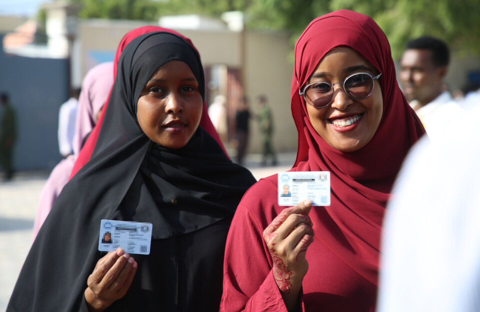 Residents cast ballots at polling stations as people in Mogadishu vote directly in local elections for the first time since 1969, marking a historic moment in Somalia's electoral process. Two women in hijabs stand in line, holding up their ID cards. One wears glasses and is smiling, while both look directly at the camera.