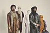 Five Tuareg men, dressed in traditional clothes and wearing turbans, are posing in front of an off-white wall and looking directly into the camera.