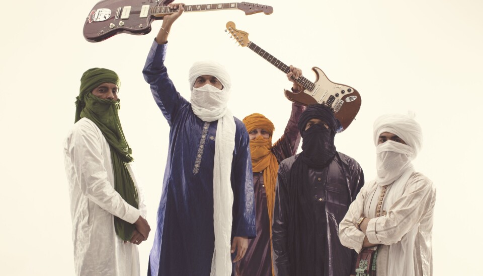 While they have built a lively community hub, the members of Imarhan are also noticing that younger generations are at risk of losing their traditions. Five men wearing traditional Tuareg attire and turbans are standing in front of a white background, looking directly into the camera. Two of them are holding electric guitars over their heads.