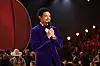 Trevor Noah speaks onstage during the 68th GRAMMY Awards, smiling while holding a mic and wearing a purple jacket.