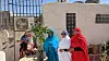 Four women wearing colourful toubs are standing around a wooden table in the courtyard of a white building, two of them are smiling and looking directly at the camera.