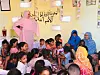 Najla Mohamed-Lamin is standing in a pink toub, over a group of young children and two women sitting by a yellow wall with Arabic writing.