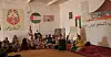 Several adults and children are sitting in a white-walled room with several Sahrawi flags and Arabic writing. A woman, Najla Mohamed-Lamin, is sitting on the floor with a microphone in hand.