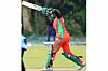 Melvin Khagoitsa, wearing a red and green Kenya cricket uniform and helmet, prepares to bat during a cricket match on a grassy field.