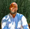 A headshot of John Mburu. He is sitting in front of a long grass plant, a slight smile on his face, wearing a light blue shirt.