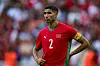 Morocco’s national football team captain, Achraf Hakimi, stands on a field, with spectators blurred behind him, wearing the Morocco national jersey, which he tugs with one hand as he looks off into the distance.