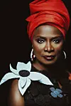 Angélique Kidjo wears a red turban and looks directly at the camera in a striking portrait promoting her new album. She also wears silver hoop earrings, dark cherry-red lipstick, and a dress with floral patterns.