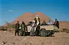 Nine men in traditional Tuareg attire, eight of whom are wearing Tuareg turbans, are gathered around a white Toyota Jeep in the desert. Behind them looms a mountain, and around them are small bushes.