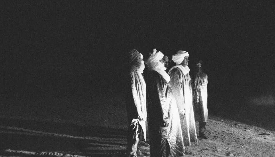 Five men in traditional Tuareg attire stand in the desert at night. A bright light is illuminating them from the front and casting shadows of their figures behind them as they look right at it.