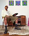 Da Capo holds a vinyl record while sitting down in a press image. Behind him is the artwork for Indigo Child II, and he wears a cream white blazer, olive shorts, and black shoes.