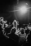 A woman with long hair is standing in the middle of a crowd and musicians, holding a microphone to her mouth and singing with one raised hand.