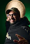A profile image of Kesivan Naidoo looking at the camera while holding a cymbal behind his head. He wears an olive NY cap and a black Azerbaijan sweater.