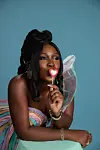 A portrait of Osas Okonyon in a colorful dress, playfully holding a red and white heart-shaped lollipop to her lips.