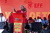Julius Malema stands at a red podium, speaking into a mic, with people behind him.