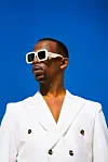 A promo picture of Zakes Bantwini against a sky blue background. He wears dark shades with a white frame and a white blazer.