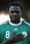 Former Super Eagles striker, Michael Eneramo, in a green and white Nigeria national football team jersey on a field with a hand over his heart, staring ahead.