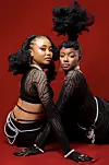 Gloria Munga (left) and Maggie Atieno (right) of We Are Nubia pose against a red background, wearing black lace outfits with pearl accessories.