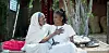 An old woman is sitting on a bench with her granddaughter, both are wearing white. The elder’s hand is placed on the younger’s chest as they look at each other.