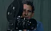 In front of a green cloth background, a middle-aged man is behind a large camera, looking critically at what it is capturing.