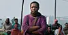 A woman in a purple dress and brown scarf stands with her arms crossed outdoors while several people sit behind her in a still from the Rwandan film 'Ben’Imana.'