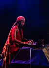 A woman with long black braids is at a DJ console, wearing a red dress and traditional Sudanese gold crescent earrings. A ghamarsis, a traditional Sudanese checkered cloth, is wrapped around her head like a long bandana.