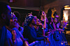 In a dark theater, several people stand in the front row at a concert, facing the stage, smiling or singing along.