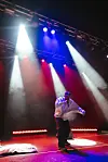 A man wearing a white hoodie and a black cap is rapping on stage, behind him, lights are shining in red, blue, and white.