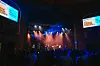 In a dark theater, people are performing on stage for a large crowd. On screens flanking the stage, a banner reads “The Sound of Sudanese Excellence.”