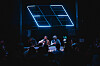 Band performing on a dark stage with blue neon light panels and crowd silhouettes in front.