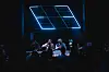 Band performing on a dark stage with blue neon light panels and crowd silhouettes in front.