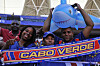 Smiling Cabo Verde fans hold a blue inflatable shark and a team scarf at a football stadium.