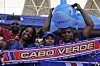 Smiling Cabo Verde fans hold a blue inflatable shark and a team scarf at a football stadium.